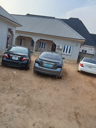 The image shows three cars parked in front of a house located in Sapele, Nigeria (latitude: 5.863, longitude: 5.703). The first car is a black Toyota Camry with the license plate "BVA 941 CF". The second car is a gray Toyota Camry with a red license plate. The third car is a white Mercedes-Benz C-Class with the license plate "BW 677 LL". The house is a single-story building with a gray roof and white walls. The yard is sandy and there are tire tracks in the sand. The house has a gray roof with metal tiles and black windows.