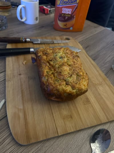 A wooden cutting board displays a loaf of golden-brown savory bread, potentially zucchini or cheese bread, positioned next to two knives. A silver spoon rests at the board's corner. Adjacent to the board is a cup with a handle, a container of Foulain coffee, and a partial view of a dark surface, possibly a couch. The overall setting suggests a casual meal or snack preparation.