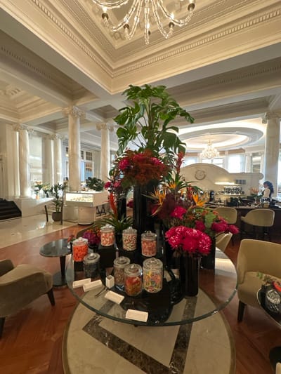 This image depicts the opulent lobby lounge of the Beau-Rivage Palace hotel in Lausanne, Switzerland. The scene is dominated by a large, round glass-topped table in the foreground, featuring a central display of nine clear glass jars filled with colorful candies and confectioneries. White paper trays and serving tongs are provided on the table, indicating a self-service arrangement. Surrounding and intermingling with the candy display are multiple black vases holding large, vibrant floral arrangements, including tall green Monstera leaves, red and pink chrysanthemums, and various exotic flowers such as Birds of Paradise.

The flooring in the immediate foreground consists of dark wood parquet, transitioning to a light-colored marble or tiled area with a dark geometric pattern beneath the central table. Two upholstered armchairs, one beige/taupe on the left and one light tan on the right, are partially visible, positioned around small dark tables, suggesting guest seating.

In the midground to the left, a grand architectural section features multiple white, fluted marble columns with ornate capitals, supporting a high ceiling with intricate white molding and cornices. A polished wooden staircase leads upwards on the far left. A modern, white display case, possibly for pastries or desserts, is visible beyond the central table, along with additional potted plants.

To the midground right, a contemporary bar or service counter with a curved white facade is visible, topped by a large, round white clock. Behind the counter, shelving stocked with bottles and glassware is apparent. Two light-colored, upholstered bar stools are positioned in front of the counter. A single individual, a staff member in a dark uniform, is partially visible behind the bar, attending to the area.

The high ceiling throughout the space is adorned with multiple elaborate crystal chandeliers, providing overhead illumination. The overall environment conveys a sense of grandeur and luxury within a meticulously maintained interior space.