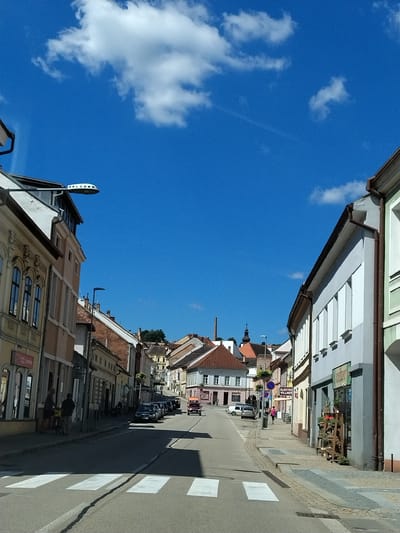 The image depicts a street scene in the city of Volyně, Czechia, under a bright blue sky with scattered white clouds. The perspective is from a low angle, looking down a cobblestone street lined with multi-story buildings. On the left, a pale yellow building with arched windows and ornate detailing is visible. To its right, a lighter colored building with a brown roof and several windows follows. The right side of the street features a building with a pale blue facade, white-framed windows, and a brown gutter system. In front of this building, a wooden display rack with plants is positioned on the sidewalk. Further down the street, several cars are parked along the side, including a black sedan, and an orange van is seen in the distance. Pedestrians are present, with two individuals standing on the sidewalk near the left-side buildings, one of whom appears to be interacting with a bicycle. Another pedestrian in a pink jacket is walking on the right-side sidewalk. A crosswalk is marked on the asphalt road in the foreground. In the background, the street curves upwards, revealing more buildings with red-tiled roofs and a distant church spire. A tall, brick industrial chimney is also visible in the background to the left. The overall impression is a sunny day in a European town.