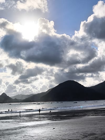 The image captures a vibrant beach scene at Selongbelanak, Indonesia, likely during late afternoon or early morning, characterized by dramatic lighting. The setting features a wide, wet sandy beach in the foreground, stretching towards the ocean where gentle waves break. In the mid-ground, numerous people are visible as dark silhouettes against the reflective water and sand. Some individuals are standing or wading in the shallow surf, while others are further out in the water, appearing to swim or play.

Beyond the immediate beach, the ocean extends to a range of dark, undulating mountains that form the backdrop. Numerous small boats or rafts are dotted across the water, adding to the local atmosphere. The sky is a prominent feature, filled with large, varied clouds. The sun, partially obscured by these clouds, creates a brilliant, intense glare in the upper left quadrant, backlighting the entire scene and creating strong contrasts. Some clouds are brightly lit, appearing white and glowing, while others are dark and heavy, suggesting a dynamic weather pattern. The overall impression is one of natural beauty and tranquil leisure, with the interplay of light and shadow on the clouds and water creating a striking visual impact. There is no visible text in the image.