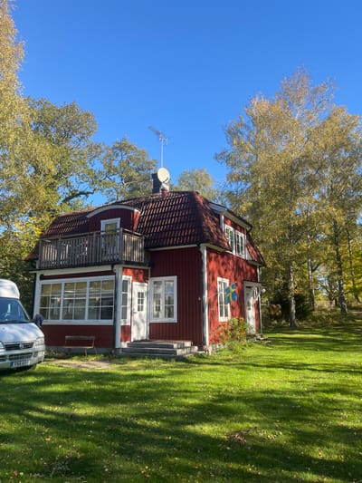 A two-story, red-painted wooden house with white window frames and door trim stands on a manicured green lawn in Västervik, Sweden. The house features a prominent gambrel-style roof with red tiles, and a second-story balcony with wooden railings is visible on the left façade. A satellite dish and a television antenna are mounted on the roof. A Swedish flag is displayed horizontally from the façade on the right side of the house, above a set of white-trimmed windows. A white Fiat Ducato camper van, bearing Swedish license plate XNA 498, is partially visible parked on the left side of the frame, adjacent to the house. A dimly visible individual is positioned near the front of the van. A small, dark-colored wooden bench sits below the ground-floor windows. The environment includes mature deciduous trees with yellow-green foliage bordering the property, indicating an autumn season. The sky is clear and blue, and direct sunlight illuminates the scene, casting distinct shadows across the grass.