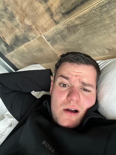 A man is lying on a bed in what appears to be a room with wooden wall paneling. He is wearing a black jacket with the word "BOSS" visible on the right sleeve. His head is resting on a white pillow. He is looking directly at the camera with a slightly furrowed brow and open mouth. The environment appears to be indoors, with a focus on the man and the immediate surroundings of the bed and wall. There is no immediately available information about the location in Pamporovo, Bulgaria.