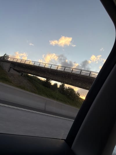 This image captures a view from within a vehicle, looking out towards a bridge crossing over a highway. The sky is a soft blue, with scattered clouds illuminated by the setting sun, creating a warm glow. The bridge itself is a concrete structure with a metal railing, and it spans across a grassy embankment dotted with trees. The highway below the bridge appears to be a multi-lane road, with a concrete barrier separating the lanes. The foreground is dominated by the interior of the car, with a dark pillar and window frame on the right side. The scene is likely captured during a drive on a highway in or near Uddevalla, Sweden, given the typical infrastructure seen in such locations.