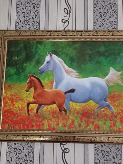 The image shows a framed painting of two horses in a field of red flowers. A large white horse is in the background, and a smaller brown horse is in the foreground. The painting is hanging on a wall with striped and patterned wallpaper. The painting is framed with a decorative gold frame. There are no people visible in the image. The scene depicted is a natural landscape with animals. There is no information in the image or its crops that would indicate the location in the city of Älimbet, Kazakhstan.