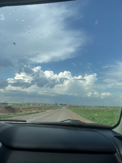 The image shows a view from inside a vehicle, likely a car or truck, looking out over a road and the surrounding environment. The sky dominates the view with a mix of blue sky and puffy white clouds. The road is straight and appears to be a highway with green fields on either side. A town with buildings and infrastructure is visible beyond the green fields. The perspective suggests the vehicle is in motion. The interior of the vehicle is visible on the right side of the frame, with the dashboard and part of the side window. The scene is outdoors, during daylight hours, with clear weather conditions but with some cloud cover. There's no evidence to suggest the city of Kitsjanovo.