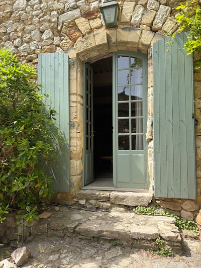 An exterior view shows a window with open light-green shutters in a building's exterior rock wall. The window is set within an arched opening. The rock wall is light brown and tan in color. Lush green vegetation is visible around the bottom of the window. A small lantern hangs to the left of the window. The location is Saint-Victor-la-Coste, France.
