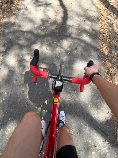 The image depicts a first-person, downward view of an individual cycling on a paved asphalt path in Gainesville, United States. The rider is mounted on a red road bicycle featuring red handlebar tape. A Garmin cycling computer is centrally mounted on the handlebars, displaying numerical data including "19:36 6.79" and "18.5". The bicycle's frame exhibits a small Swiss flag decal and a yellow sticker with "BikeReg.com" text. The rider's bare legs, clad in black cycling shorts, are visible, along with white cycling shoes (Giro brand visible on the right shoe) and white socks. The rider's right hand, wearing a black wristband and a pink/white braided bracelet, grips the right handlebar drop. The paved path is partially covered with scattered dry, fallen leaves, primarily brown and yellow. Overlying the path are distinct shadows cast by unseen overhead trees, indicating a sunny, outdoor environment. The path's edges are lined with additional fallen leaves and natural ground cover.