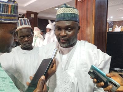 The image depicts a scene of a man being interviewed, likely in a public building. The man, wearing a white long-sleeved robe and a blue and white patterned cap, is being interviewed by a person holding a smartphone. To the man's left, a second man, also wearing a white robe and a cap, is holding a smartphone. The scene takes place inside a building with wooden paneling on the walls. The image appears to have been taken in Kaltungo, Nigeria.