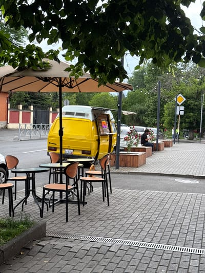 A yellow food truck is parked on a paved street next to a sidewalk cafe. The food truck has its side door open, and a neon sign is visible inside. Several outdoor tables and chairs are set up on the sidewalk in front of the cafe. A man is sitting on a wooden bench near a planter filled with red roses. Trees and string lights are visible in the background. The scene appears to be in a city, with buildings and a road visible. The license plate of the food truck has the text "AVE BUS".