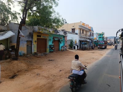 The image depicts a street scene in Kothacheruvu, India. In the foreground, a man is riding a motorcycle on the right side of the road. The road is partially paved, with a dirt shoulder on the left. Along the left side of the street are several buildings, some with closed storefronts. One building features a sign in Telugu advertising animal feed, including cotton seed cake from S.L.N.S. and S.K.M 777 brands. Other buildings have signs for "Sri Kiran General Store" and "Suresh". In the background, a truck with "Subhan Allah" written on it is visible, along with other vehicles and buildings. A tree is present near the storefronts on the left.