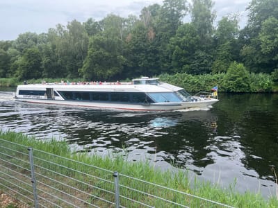 A white tour boat named "BELVEDERE" is traveling on a dark body of water, likely a river or canal. The boat has a modern design with large, tinted windows and a white upper deck. On the upper deck, numerous people are visible, standing and seated, enjoying the ride and the scenery. The boat is emitting a wake, indicating it is in motion.
The scene is set against a backdrop of lush green trees and vegetation lining the water's edge. The trees are dense and form a natural barrier along the bank. The sky is overcast with a cloudy, muted light, suggesting it is daytime, likely late morning or afternoon. A metal fence with vertical and horizontal bars runs along the foreground, separating the viewer from the water and the grassy bank. The water is dark and reflects the overcast sky and the surrounding trees, creating a textured surface.
On the stern of the boat, a flag with red, white, and blue colors is visible, along with a yellow and blue striped flag. Text on the side of the boat reads "SCHIFFFAHRT IN POTSDAM.de". The overall atmosphere appears calm and leisurely, typical of a scenic boat tour. The location is Teltow, Germany.