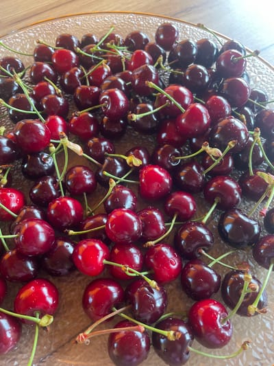 A high-angle, close-up photograph displays a collection of approximately 50-70 fresh cherries arranged within a clear, textured glass plate. The cherries exhibit varying shades of red, ranging from a vibrant ruby red to a deeper, almost black-red hue. Numerous small water droplets are visible on the skin of many cherries, suggesting recent washing or freshness. Most cherries retain their green stems, often clustered, with some showing light brown calyx remnants at their attachment points. The glass plate features a raised, faceted, or dimpled pattern on its surface, causing light refraction. The rim of the plate, visible at the top and right edges, appears to be made of glass with a slight amber or golden tint due to reflections. The plate rests on a smooth, polished wooden surface, visible as a warm brown background with subtle grain patterns. The scene is evenly illuminated, producing highlights on the wet, glossy cherry skins. There are no people, explicit actions, or interactions depicted.