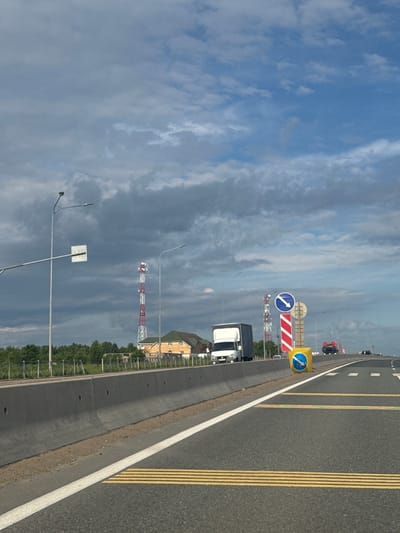 The image depicts a multi-lane highway under a partially cloudy blue sky. The road features clear markings, including a white solid line separating the shoulder from the main lanes, double solid yellow lines on the far-right lane, and dashed white lines for inner lanes.

Several vehicles are visible, including a white semi-truck in the mid-ground and a red truck further down the road, suggesting ongoing traffic. Road signs are prominent: a blue circular sign with a white arrow pointing straight ahead (or slightly right) is seen on the left side of the road, and immediately next to it, a red and white striped rectangular sign, possibly indicating a lane closure or hazard. Another blue circular sign with an arrow pointing straight ahead (or slightly left) is encased in a yellow square frame further down the highway, acting as a directional sign. A smaller, similar blue arrow sign is also visible in the distance. A white square sign on a pole is on the far left, but its content is illegible.

The highway is bordered by a concrete barrier on the left. Tall streetlights are spaced along the road. In the background, beyond a line of green trees, a large red and white striped communication or utility tower stands out, with another similar tower visible further away. A light-colored building with a dark roof can be seen behind the trees and the first tower. The scene appears to be set during daytime, with ample natural light. The context provided indicates the location is Beloborodovo, Russia.
