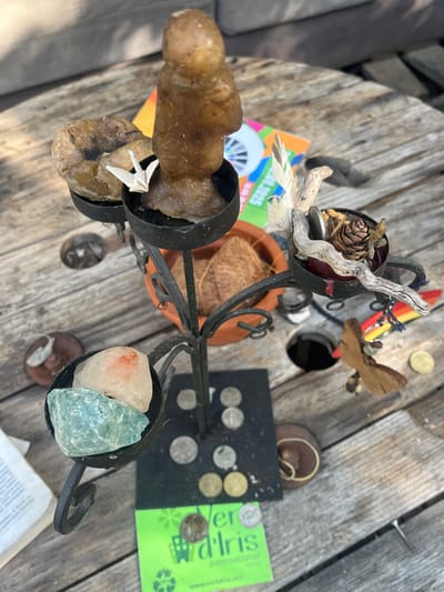 The scene is an overhead view of a collection of diverse objects displayed on a weathered wooden plank surface, characteristic of an outdoor table, located in Sint-Pieters-Leeuw, Belgium.
At the center is a black, multi-tiered wrought-iron stand with four candle holders, utilized for displaying various items. The top-left holder contains a large, irregular light brown stone or mineral specimen. The top-center holder features a brown, amorphous figure, possibly crafted from wax or stone, with a small white origami paper crane placed at its base. The top-right holder is filled with natural elements including two white feathers, several pieces of light-colored driftwood, a single pinecone, and a few small, dull metallic objects, possibly coins or pebbles. The bottom-left holder contains two raw mineral specimens: one predominantly light pink/orange and the other turquoise/light green.
Scattered on the wooden surface around the base of the stand are several European coins of various denominations and metals (silver-colored and gold-colored). A bright green business card or flyer for "Ver d'Iris International" with a recycling symbol is visible near the stand's base, partially covered by one of the coins.
In the background, behind the stand, a terracotta pot holds a brown, desiccated coconut husk. Further back, a colorful book or magazine with "2023" visible text is partially obscured. To the right of the stand, a small, multi-colored surfboard-shaped charm hangs from a string, and a wooden bee or insect figurine is positioned nearby, alongside an additional gold-colored coin and a dark, ring-shaped object. To the left, a page from a book or document with visible text lies open. The environment is illuminated by natural light, casting soft shadows. No individuals are present in the image.