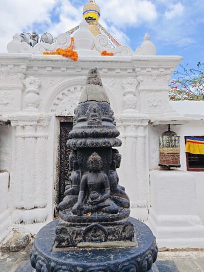 In Gokarneshwar, Nepal, a black stone sculpture of Buddha figures sits on a pedestal in front of a white temple. The sculpture is detailed, with each Buddha seated in a meditative pose. The temple behind it has ornate white plasterwork and a large, dark wooden door. Prayer flags and orange marigold garlands adorn the temple's roofline, leading up to a golden stupa in the background. A prayer wheel hangs on the right side of the temple wall, next to a window draped with a yellow and blue cloth. The sky above is partly cloudy.