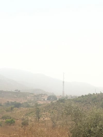 The scene depicts a panoramic, hazy view of a rural landscape in Kwakwa Laji, Nigeria. The upper half of the image is dominated by a diffused, bright white sky indicating significant atmospheric haze, which reduces visibility of the distant background. Faint, obscured mountain ranges or hills are discernible through the haze along the horizon. In the midground, a tall, slender, lattice-structure telecommunications tower stands prominently on a gentle slope towards the right of the frame's center. Scattered residential or agricultural structures are indistinctly visible near the base of this tower and further left. The terrain features a mix of sparse, dry, brown grasses and patches of green and deciduous trees and shrubs. The immediate foreground is characterized by dense, dry, golden-brown grasses and low-lying scrub. No individuals, animals, or specific activities are visible within the frame. The overall environment appears dry and expansive.