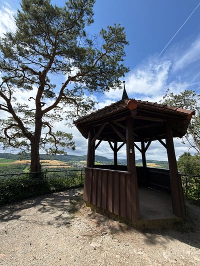 A wooden gazebo with a dark brown, tiled roof sits on a gravelly overlook, offering a view of a rolling, green landscape under a blue sky with scattered clouds and a distant contrail. The gazebo is octagonal, with vertical wooden planks forming its lower walls and open upper sections supported by wooden beams. A metal fence runs along the edge of the overlook, separating it from the scenic vista. To the left of the gazebo, a large pine tree with a gnarled trunk and sparse foliage casts a shadow on the gravel. The landscape beyond features a patchwork of fields, forests, and a small village in the distance. The scene appears to be a viewpoint in or near Heiligenstadt i. OFr., Germany, possibly a park or natural area. There are no people or animals visible in the image.