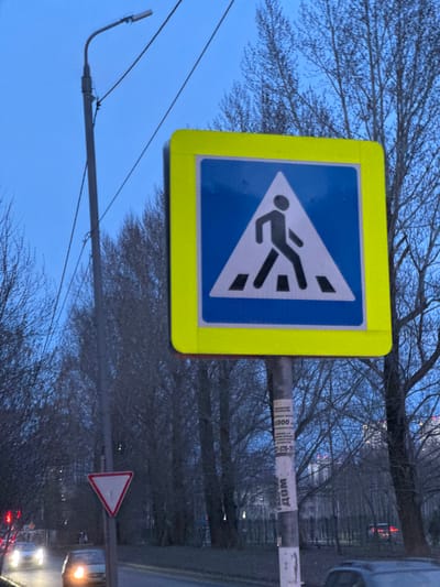 The image shows a pedestrian crossing sign in Matiushino, Russia. The sign is a yellow square with a blue triangle inside, depicting a person walking across a crosswalk. The sign is mounted on a pole with advertisements attached. In the background are trees without leaves, a road with cars, and a yield sign. The sky is a muted blue color.