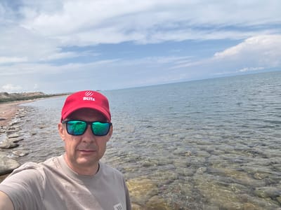 A man with a red baseball cap and sunglasses takes a selfie by the shore of Issyk-Kul Lake in Tong, Kyrgyzstan. The man is wearing a light brown t-shirt. The lake's water is clear, revealing pebbles on the lakebed. The shoreline is rocky with some dirt and sparse vegetation. The sky is partly cloudy with blue sky visible. In the background, the land slopes upwards, and some structures are visible on the hill.