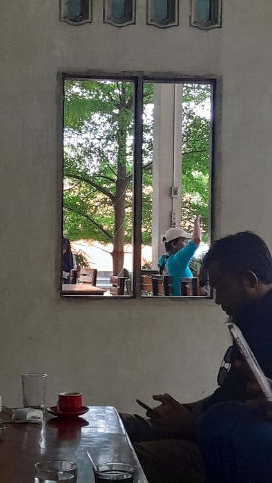 The image displays an indoor perspective looking through a framed window onto an outdoor setting, located in Keudealue Ie Puteh, Indonesia. In the foreground, positioned to the right, a person with dark hair and a beard, dressed in a dark shirt and dark pants, is seated. They are observed holding a smartphone in their left hand and a larger mobile device in a white case in their right hand, both being actively used. A dark, reflective wooden table occupies the lower left and center of the frame. On this table, a clear glass tumbler, a red cup on a matching red saucer, and two smaller clear glasses are present; one small glass contains a dark liquid with a spoon, while the other appears empty. The interior wall is a light, textured surface, featuring three decorative, blue-tinted ventilation blocks above the window.
The window itself has a dark frame and is split vertically into two panes. Several small brown pots are arranged along the window sill. Through the window, an outdoor environment is visible. A second individual, wearing a light blue shirt and a white baseball cap, stands to the right of the window's vertical divider, with their left arm raised in a gesturing or waving motion. Green trees with visible branches and foliage dominate the upper and left portions of the outdoor view, while a light-colored architectural pillar stands behind the second person. The scene is illuminated by natural daylight.