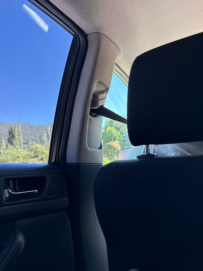 The image is taken from inside a car, looking out the passenger window. The view outside shows a clear blue sky, mountains in the distance, and green trees and foliage. The interior of the car is partially visible, including a black headrest of a seat in the foreground, the car's door panel with a chrome door handle, and a section of the car's headliner. A seatbelt is also visible. The image does not contain any information about the location being Ασπροβάλτα, Greece.