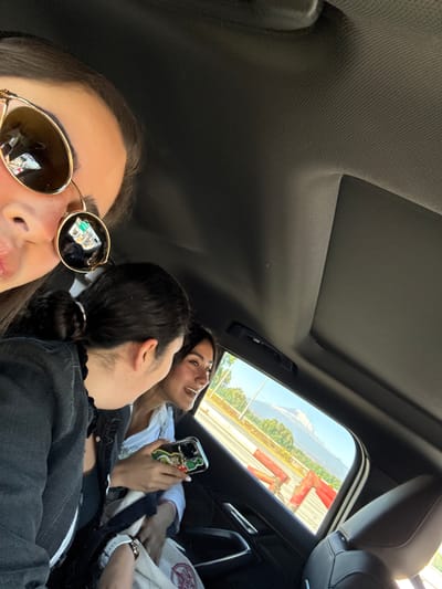 The image captures the interior of a vehicle with three occupants, situated in or near Santa María Moyotzingo, Mexico, based on the external landscape.
In the immediate foreground, the upper left portion of a person's face is visible, wearing large, round, gold-rimmed sunglasses with dark lenses. Reflections in the sunglass lenses show ambient light and indistinct interior details.
To the right of the foreground person, a second individual is seen from a side and rear perspective. This person has dark hair pulled back into a bun or ponytail and wears a dark upper garment. A white-banded watch is visible on their left wrist. They are turned towards a third individual, suggesting interaction.
The third individual is positioned further back on the passenger side. This person has dark hair, wears a light-colored top, and is smiling while looking at the second individual. They hold a smartphone with a colorful, character-themed case, specifically identifiable as Mickey Mouse, in their right hand.
The vehicle's interior features a dark headliner and a visible sunroof opening. A dark, upholstered car seat headrest is partially visible on the far right of the frame.
Through the passenger side window, an external scene unfolds. A road with a yellow dividing line is visible, bordered by red and orange plastic traffic barriers. Beyond these barriers, green trees and vegetation are present. In the distant background, a large, prominent snow-capped mountain or volcano is clearly visible under a clear blue sky, consistent with views of Popocatépetl or Iztaccíhuatl from the Santa María Moyotzingo region.