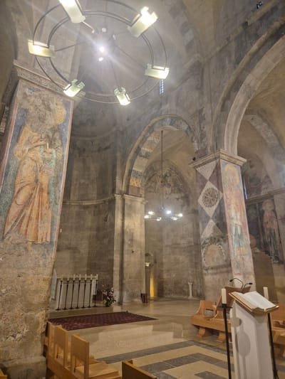 The image shows the interior of the Church of the Ark of the Covenant in Abu Gosh, Israel. The church features ancient stone architecture with rounded arches and vaulted ceilings. The walls and pillars are adorned with faded frescoes depicting religious figures and geometric patterns. A chandelier with multiple lights hangs from the ceiling. In the foreground, wooden pews are arranged in rows. An altar with candelabra and a floral arrangement is visible in the middle ground, along with a red rug. A lectern with an open book and candles stands to the right. The overall impression is one of historical and spiritual significance, with the ancient artwork and architecture contributing to a solemn atmosphere.