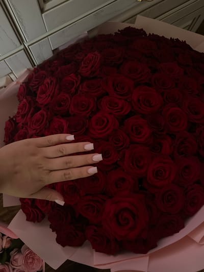 A hand with light pink, square-shaped nails is shown resting on a large bouquet of deep red roses. The roses are tightly packed, creating a voluminous display. The bouquet is wrapped in pale pink paper, with some of the paper peeking out from beneath the blooms. To the left of the bouquet, a section of white tiled wall with a distressed wood-like texture is visible. In the lower right corner, a small arrangement of pale pink roses can be seen. The lighting suggests an indoor setting, possibly during the evening or in a dimly lit room. The overall mood is romantic and luxurious, with the rich red of the roses and the elegant presentation. No text is visible in the image.