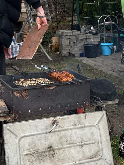 The image depicts a barbecue scene, possibly in a backyard or garden in Dārzciems, Latvia. The focus is on a large, black metal barbecue grill with a light grey lid. Inside, meat is cooking on skewers and in a wire basket. Smoke emanates from the grill. An individual wearing a black jacket and a watch holds a reddish-brown slab or tile near the grill. Other items in the background include a blue bucket, black pots, and stacks of grey paving stones. There are indications of vegetation and garden tools. The ground appears to be a mix of gravel, soil, and some grass.