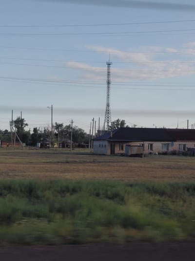 The image depicts a rural or semi-rural landscape with a low-rise building and a tall transmission tower under a pale sky with scattered clouds. The building appears to be a long, single-story structure with a dark roof and light-colored brick walls. It has several windows. A smaller, corrugated metal structure is situated in front of the building. The transmission tower, which is tall and lattice-like, is visible in the background to the left of the building. Several utility poles with wires stretch across the frame. The foreground shows a field of dry grass, and a blurred strip of green vegetation runs across the bottom of the image, suggesting the photo was taken from a moving vehicle. The overall scene is sparsely populated with hints of infrastructure in a natural setting. The location is identified as the city of Молодёжный, Kazakhstan.