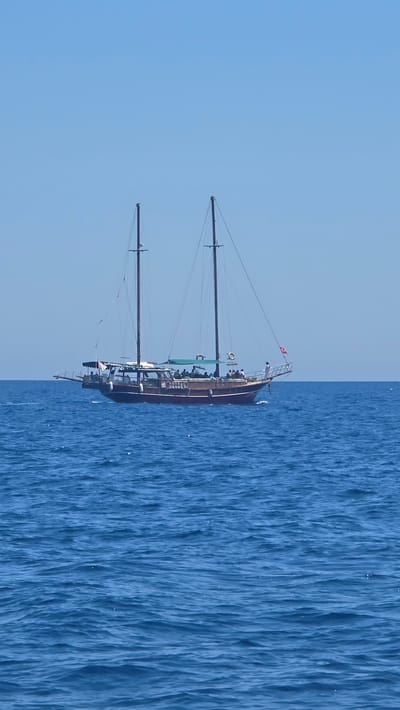 A wooden sailboat with two masts is sailing on a bright blue ocean under a clear sky. The boat has a dark wooden hull and a white deck with several people visible on it. Some people are standing, while others are seated under a canopy. The boat is flying a Turkish flag at its bow. The sea is calm with gentle ripples. The image appears to be taken from another boat or from shore, looking out towards the sailing vessel.