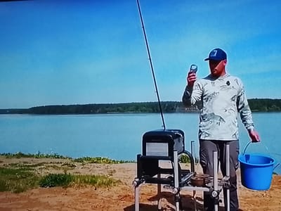 A man in a camouflage long-sleeved shirt and a blue baseball cap stands on a sandy bank next to a body of water, likely a lake or river. He is holding a small, silver object in his right hand and a blue bucket in his left. A fishing rod stands upright behind him. In front of the man is a black fishing seat box with metal legs. The background shows a wide expanse of light blue water, with a tree-lined shore on the opposite side of the water. The sky is clear and bright blue, suggesting a sunny day. The overall scene appears to be a fishing excursion in an outdoor, natural setting. The time of day appears to be daytime, given the bright sunlight and clear sky.