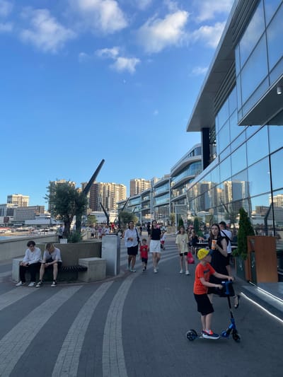 A group of young people are gathered on a paved walkway in front of a modern building with a glass facade. The building reflects the sky and surrounding urban landscape, which includes tall buildings and trees. The sky is clear blue with a few wispy clouds and a faint crescent moon. The scene appears to be set during the daytime, likely late afternoon or early evening, given the lighting.

Several individuals are visible, some standing and others sitting. One young person, wearing an orange shirt and a yellow cap, is standing on a blue scooter. Another young person in a black shirt holds an orange cup. The group seems to be casually interacting, with some appearing to be conversing or observing their surroundings. Two individuals are sitting on a bench in the foreground, also in casual attire.

The overall atmosphere seems relaxed and social. The paving stones of the walkway have a distinctive striped pattern. The architectural style of the building suggests a contemporary urban environment. The location is identified as Novomyshastovskaya, Russia. No visible text is present in the image.