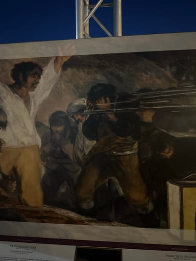 The image displays a reproduction of Francisco de Goya's painting, "The Third of May 1808 in Madrid." In the foreground, a man in a white shirt and brown pants, with his arms raised, faces a firing squad that is not fully visible. To his right, a group of men with grim expressions are preparing to be executed, some covering their faces. In the background, there is a dark, indistinct landscape. Below the painting, text identifies the artwork and the artist, Francisco de Goya, and indicates it is a "SPANISH PAINTING." There are also some Spanish inscriptions, one of which reads "FRANCISCO DE GOYA" and "El 3 de mayo de 1808 en Madrid." The reproduction is part of an exhibition displayed outdoors, indicated by a metal structure and the dark sky above. The text suggests this exhibition is located in Mazarrón, Spain.