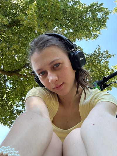 The media file is a low-angle selfie of a young woman with headphones on, looking up at the camera. She is wearing a yellow t-shirt. Her hair is brown and pulled back. The background consists of green trees and a clear blue sky, suggesting an outdoor setting during the daytime. Sunlight filters through the leaves, creating dappled lighting. The woman's expression is neutral. The scene appears to be a casual moment, possibly while relaxing or enjoying an outdoor activity. There are no visible text elements in the image. The metadata indicates the location is Erlenbach am Main, Germany.