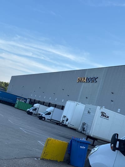The image depicts a wide shot of a loading dock area with a large industrial building in the background. The building features the prominent signage "DYNALOGIC" in large, golden letters with a blue outline. The facade of the building is made of corrugated metal and has several loading bays.
In front of the building, several white trucks and trailers are parked, some with their doors open, suggesting active logistics operations. One trailer has "Becx verhuur" branding. A blue shipping container with "READYPAL" branding is also visible. The ground is paved asphalt, marked with white lines indicating parking spaces.
In the foreground, a bright yellow concrete barrier is present, along with a blue recycling bin labeled "EUROKRAFT." A portion of a white vehicle, likely another truck or van, is visible in the lower right corner. The sky above is a clear blue with scattered, wispy white clouds, suggesting daylight conditions.
The provided context places this scene in the city of Boom, Belgium. This indicates a commercial or industrial zone within the city, likely a distribution or logistics hub. There are no people clearly visible in the image, but the presence of vehicles and the loading bay infrastructure implies human activity related to transportation and warehousing.