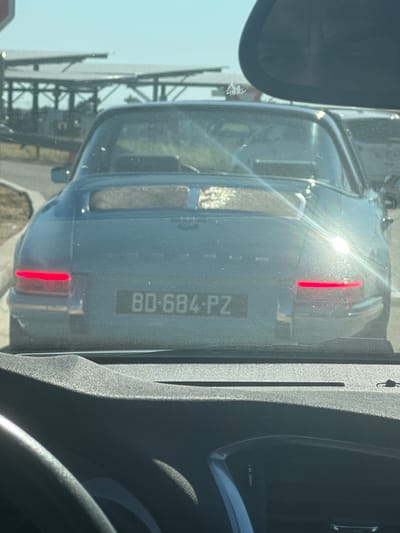 The image shows the rear of a light blue classic car, likely a Porsche, with a license plate reading "BD-684-PZ". The car is parked in an outdoor setting, possibly a parking lot or roadside, with solar panels visible in the background. The scene is viewed from inside another vehicle, indicated by the dashboard and steering wheel in the foreground. The lighting suggests it is daytime, with sunlight creating lens flares and reflections on the car's surface. The location is identified as Colombier-Saugnieu, France. There are no people clearly visible in the image. The overall scene is a snapshot of a car in a public space.