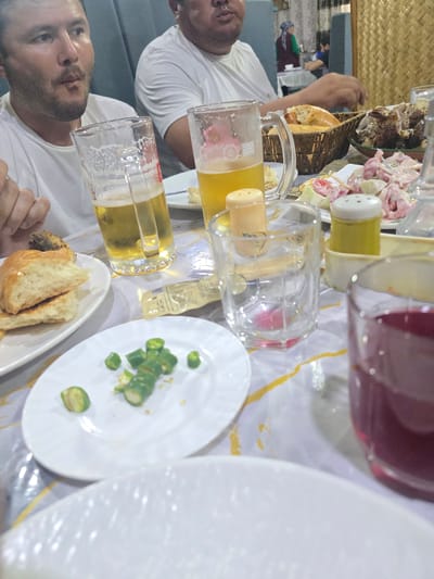 This is a close-up, tilted shot of a table laden with food and drinks, likely during a meal. Two individuals, seen from the chest up, are present. They are both wearing white t-shirts and appear to be engaged in conversation or enjoying their meal. One man on the left is looking towards the camera with a neutral expression. The other man, on the right, is looking slightly away.

On the table, there are several glasses, some containing yellow liquid, presumably beer. There's also a glass with a darker red liquid. A white plate holds sliced green chilies, and another white plate contains pieces of bread and what looks like a small portion of meat. A woven basket holding bread is also visible. In the background, there's a dish with some kind of meat, and a pink salad.

The setting appears to be indoors, possibly a restaurant or a home. The lighting suggests it could be daytime, or at least well-lit indoors. The overall mood seems relaxed and convivial, as is typical of a shared meal. There is no visible text in the image. The scene is set in Guzar, Uzbekistan.