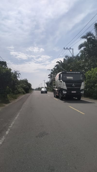 This is a medium-angle, eye-level, static shot of a two-lane asphalt road in Kruengseupeng, Indonesia, on a cloudy day. The road runs straight into the distance. On the right side of the road, a white cement mixer truck with the text "GAJAH PUTEH BETON" on its side and license plate "B 9685 XJY" is driving away from the camera. On the left side of the road, a dark-colored sedan is driving towards the camera. The landscape on both sides of the road is lined with palm trees and other dense green foliage. Utility poles with power lines are visible along the roadside. The sky is partly cloudy with a bright, diffused light.