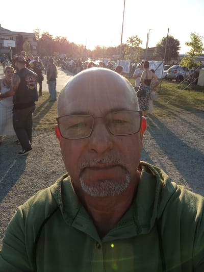 An outdoor, close-up selfie captures a bald, middle-aged Caucasian man wearing glasses and a green hooded shirt. He has a goatee and appears to be facing the camera directly. The background shows a gathering of people and motorcycles in what seems to be an open area, possibly a parking lot or field, with trees and buildings in the distance. The sun is setting, casting long shadows and creating a warm, golden light. The scene suggests a social event, perhaps a motorcycle rally or festival, taking place in Arbia, Italy, given the context provided.