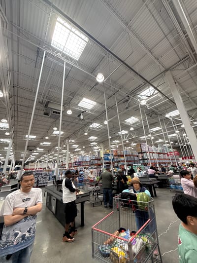 The image depicts the interior of a large warehouse-style retail store, likely a Costco, located in the City of Yonkers, United States of America. The scene is populated with numerous shoppers and employees. The architecture features a high, corrugated metal ceiling with exposed beams and numerous fluorescent lights and skylights. Racks of merchandise, including boxes of food and household items, are visible throughout the store.

In the foreground, a man wearing a white graphic t-shirt and jeans stands with his arms crossed, looking towards the camera. To his right, another man in a white and black t-shirt and black shorts stands at a checkout counter. In the middle ground, several people are pushing shopping carts and interacting at checkout stations. One woman in the middle ground is wearing a wide-brimmed hat and looking at a watermelon in her shopping cart. Further back, more shoppers are visible browsing merchandise and moving through the store. The overall atmosphere appears to be one of a busy shopping day.