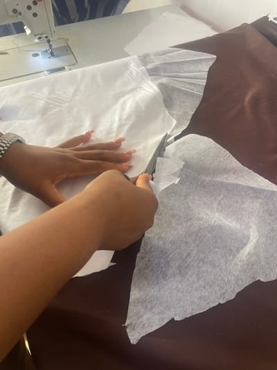 The image captures a high-angle view of a person's hands engaged in textile work, likely in a workshop or crafting setting in Dushepe, Nigeria. The primary activity shown is cutting fabric. A pair of hands is using dark-handled scissors to precisely cut a white, translucent, non-woven material. This white fabric, which appears to be lightweight and has a somewhat fibrous texture, is laid out over a darker, brown fabric.
The left hand, adorned with long, light-colored manicured nails and a metallic link-style watch on the wrist, steadies the fabric. The right hand holds the scissors, actively snipping through the material, creating small, irregular offcuts. In the lower-left corner of the frame, a white industrial-style sewing machine is partially visible, with a light illuminating its needle area and a blue thread threaded through it, suggesting the subsequent stage of the textile project. The scene is brightly lit, indicative of daytime or a well-lit indoor environment. There is no visible text in the image, and no specific emotions are discernible from the hands and objects.
