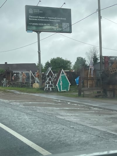 The image depicts a street scene in Zavidovo, Russia, likely taken on a rainy day based on the wet asphalt. A large advertising billboard is visible to the left, promoting a business class real estate venture called "Forest Shore." The sign displays the phone number +7 (495) 023-56-76 and indicates the location is 12 km ahead. In the background, a cluster of small, ornamented wooden structures is present on the roadside, possibly decorative or part of a local attraction. Overhead utility lines are visible. A partial view of a vehicle is seen in the lower right corner of the image.