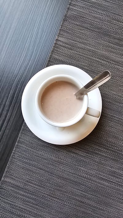 A close-up, overhead shot shows a white mug of hot chocolate on a white saucer, placed on a textured grey placemat. A silver spoon rests inside the mug, partially submerged in the beverage. To the left of the placemat is a dark grey wooden surface. The scene is indoors, likely in Alhama de Aragón, Spain, suggesting a moment of relaxation or a break.