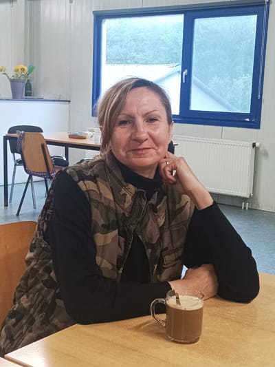 A middle-aged Caucasian woman with blonde-brown hair, wearing a camouflage vest over a black long-sleeved shirt, is seated at a light wood table indoors. Her right hand is resting on her chin, with a gold ring on her index finger. On the table in front of her is a clear glass mug filled with a light brown beverage, likely coffee or hot chocolate, with a spoon resting inside. The background shows a window with a blue frame overlooking trees, a white radiator, and a wall. In the upper left corner, a small potted plant with sunflowers is visible on a shelf. The overall environment appears to be a casual indoor setting, possibly a break room or cafeteria. The location is identified as Sexbierum, Netherlands.