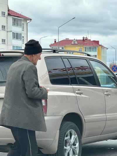 A senior man wearing a black beanie, grey jacket, and dark pants is examining a light grey SUV parked on a street in Beloyarsky, Russia. The man is looking at the rear of the vehicle and holding a small white object, possibly a pen or marker. The vehicle is parked on a city street with buildings visible in the background, including a yellow and red building with a steeply pitched roof and a white building with multiple windows. A "no stopping" traffic sign is also visible. The sky is overcast.
