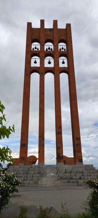 The image depicts a tall, three-part structure in the city of Araks, Armenia. It is constructed of reddish-brown stone with a pattern of darker blocks distributed throughout. The structure is composed of three parallel columns, connected by arched openings. Within the upper arches, black bells are suspended. The structure rests upon a wide, tiered stone base with two cannon replicas on top, near the base of the columns. The background consists of a cloudy sky with patches of blue visible through the structure's openings. In the foreground, there is vegetation, including flowers, partially obscuring the view of the base.