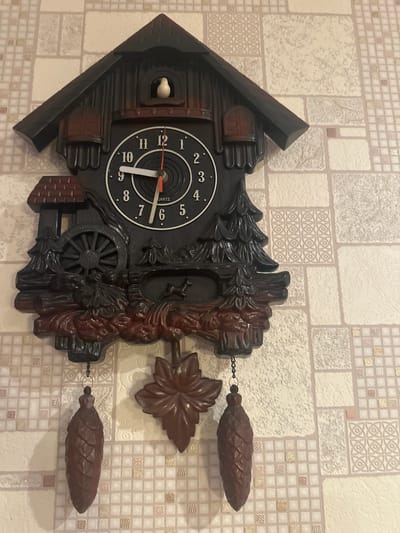 A cuckoo clock is hanging on a tiled wall. The clock has a dark brown, carved wooden casing that resembles a small house with a pointed roof. The clock face is black with white numbers and hands. The hour hand points to 10, and the minute hand points to 2. A small white bird is visible in a window above the clock face. The clock has two decorative pinecone-shaped pendulums hanging from chains. The wall is covered in a cream-colored tile with a repeating pattern of squares and textured blocks. The scene appears to be indoors, likely in a home or public space, given the presence of the clock and the wall decor. There is no indication of the time of day, weather, or emotions. The visible text on the clock face reads "QUARTZ". The location context provided is Пярэжырскі сельскі Савет, Belarus.