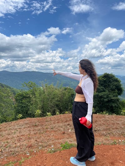 In Nao Sẻ, Vietnam, a woman stands on a reddish-brown dirt hill, pointing towards a vast expanse of lush green mountains and rolling hills under a bright blue sky dotted with fluffy white clouds. She has long, dark, curly hair and wears a brown bikini top, a white long-sleeved shrug, and black loose-fitting pants. She is holding a red water bottle in her right hand. The terrain in front of her appears to be recently cleared, with patches of dry grass and scattered debris. The background features dense vegetation, including bamboo and broad-leafed trees, receding into the distant, mist-covered mountain ranges. The overall scene suggests an outdoor activity like hiking or exploring in a natural, scenic setting.